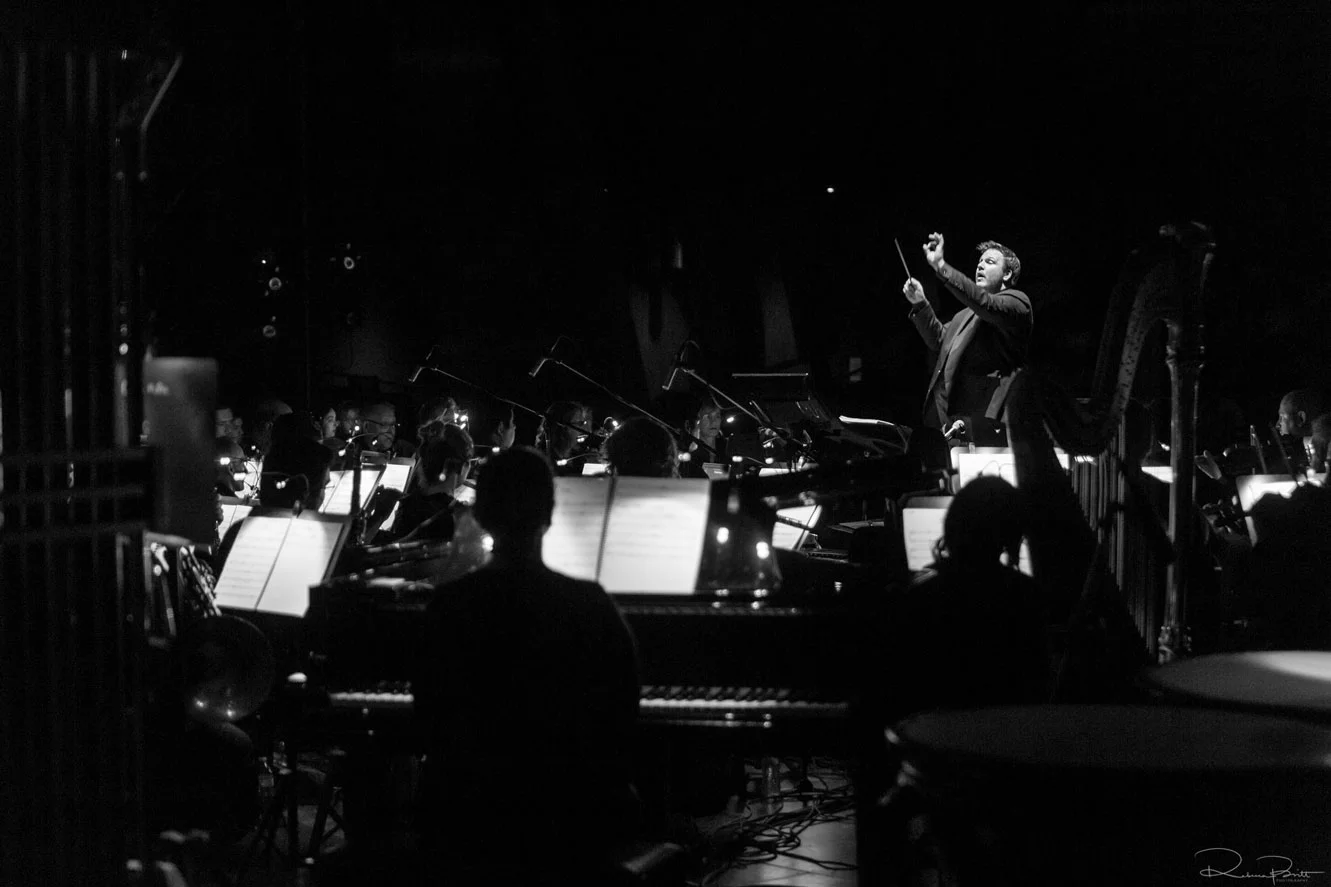A conductor leading an orchestra in a dark concert hall, in black and white