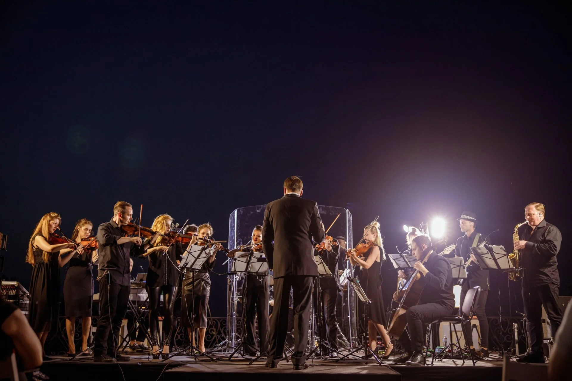 An orchestra performing outdoors at night with the conductor facing the musicians